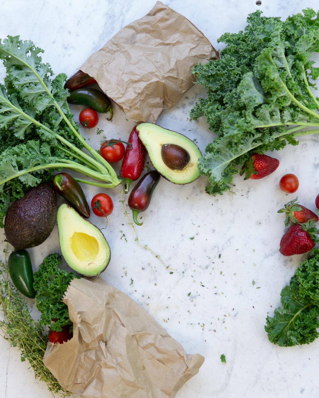 Fresh colorful vegetables including kale, avocados, tomatoes, and strawberries arranged on marble surface